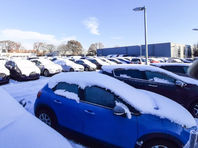 Auf einem großen Parkplatz stehen mehrere neue Wagen die mit Schnee bedeckt sind zum Verkauf