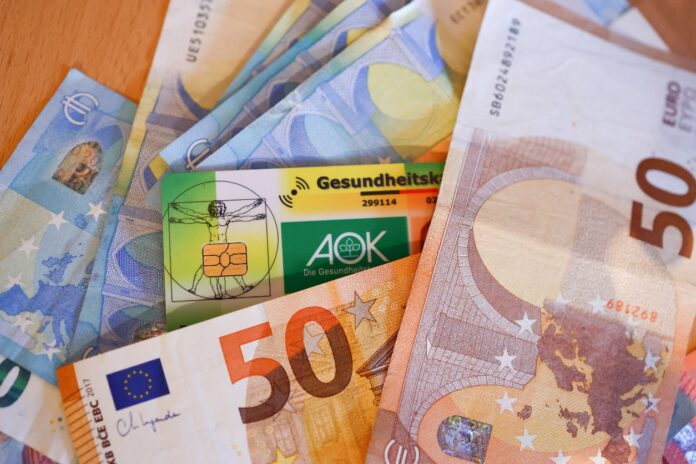 Zwischen mehreren Geldscheinen, die sich auf einem Tisch befinden, liegt eine Krankenkassenkarte. Es sind mehrere 50-Euro- und 20-Euro-Scheine zu sehen. Die Krankenkassenkarte ist von der AOK.