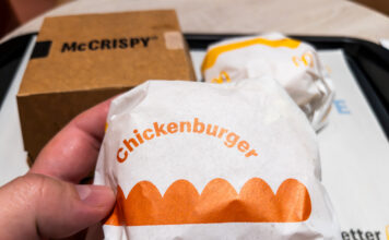 Eine Hand hält einen Chickenburger von McDonalds in die Kamera. Im Hintergrund steht auf dem Tisch das Tablet mit einem McCrispy und einem weiteren Burger.