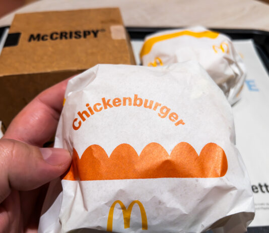 Eine Hand hält einen Chickenburger von McDonalds in die Kamera. Im Hintergrund steht auf dem Tisch das Tablet mit einem McCrispy und einem weiteren Burger.