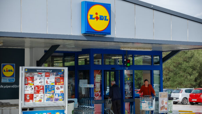 Der Eingang einer Lidl-Filiale mit geöffneten Türen und einem Schaukasten mit den aktuellen Angeboten der Woche. Im Hintergrund ist der Parkplatz inklusive mehrerer parkender Autos zu sehen.