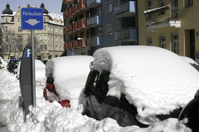 Autos, die im Winter auf dem Parkplatz einer Straße stehen. Die Autos sind von einer dicken Schneeschicht bedeckt. Neben den Autos steht ein Parkscheinautomat. Im Hintergrund Mehrparteienhäuser in verschiedenen Farben.