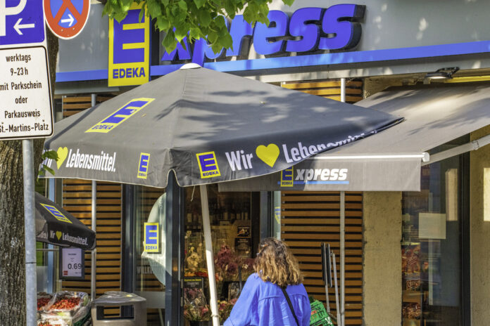Eine Filiale der Supermarkt-Kette Edeka mitten in einer Stadt. Es ist ein heller, sonniger Tag und eine Kundin steht direkt vor dem Eingang, bereit, einzutreten und einzukaufen.