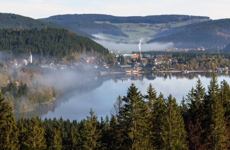 Ein großer, blauer See befindet sich in Baden-Württemberg. Er liegt zwischen einem Wald, der sich fast komplett um ihn herum erstreckt. Auf der gegenüberliegenden Seite vom Wald befindet sich eine Siedlung.