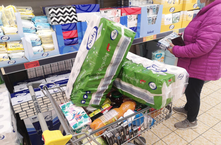 Eine Frau steht vor einem Regal mit Toilettenpapier und Kosmetiktüchern. Im Vordergrund steht ein Einkaufswagen mit viel Toilettenpapier.