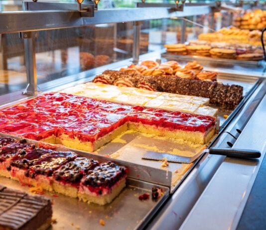 In der Theke einer Bäckerei stehen verschiedene frisch gebackene Kuchenstücke zur Auswahl für den Verkauf. Im Hintergrund sind Sitzgelegenheiten für Kunden zu sehen.
