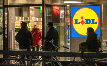 Mehrere Kunden betreten eine Filiale vom Discounter Lidl, um ihre Einkäufe zu erledigen. Es ist Abend, die Personen sind nur schattiert zu erkennen. Das Lidl-Symbol leuchtet am Eingang.
