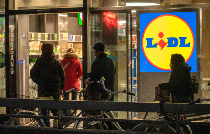 Mehrere Kunden betreten eine Filiale des Discounters Lidl, um ihre Einkäufe zu erledigen. Es ist Abend, die Personen sind nur von hinten zu erkennen. Das Lidl-Symbol leuchtet am Eingang.