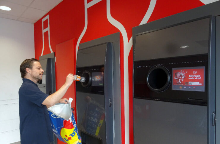 Ein Mann wirft eine Pfandflasche in den Automaten, die er aus einer Plastiktüte genommen hat. Neben dem Pfandautomaten steht zu beiden Seiten jeweils noch ein Pfandautomat.