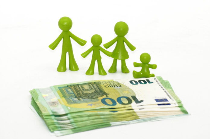 Eine vierköpfige Familie mit Vater, Mutter und zwei Kindern, bestehend aus grünen Plastik-Figuren, steht hinter einem kleinen Stapel von 100-Euro-Scheinen, die leicht aufgefächert auf einer weißen Oberfläche liegen.
