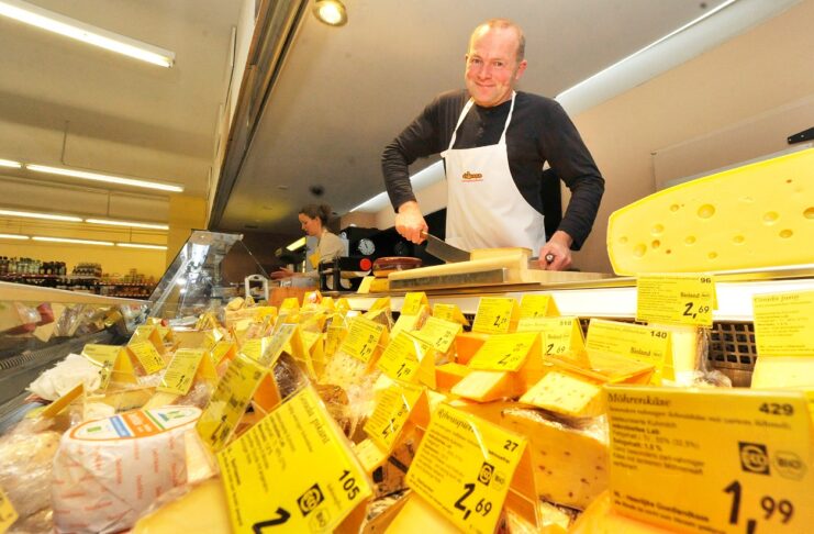 Ein Händler steht auf seinem Wagen und verkauft Käse auf dem Markt. In der Auslage befinden sich verschiedene Schnittkäse, Stückkäse, Camembert und Frischkäse.
