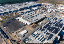 Ein Luftbild eines großen, abgelegenen Werks/einer industriellen Produktionsstätte mit mehreren Gebäuden, Nutzfahrzeugen und Parkplätzen. Im Hintergrund schließt eine leere Feldfläche die gewaltige industrielle Anlage an.