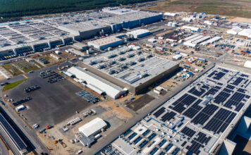 Ein Luftbild eines großen, abgelegenen Werks/einer industriellen Produktionsstätte mit mehreren Gebäuden, Nutzfahrzeugen und Parkplätzen. Im Hintergrund schließt eine leere Feldfläche die gewaltige industrielle Anlage an.