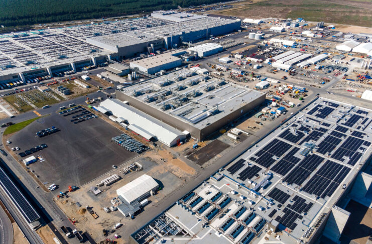 Luftbild eines großen, abgelegenen Werks/einer industriellen Produktionsstätte mit mehreren Gebäuden, Nutzfahrzeugen und Parkplätzen. Im Hintergrund schließt eine leere Feldfläche an.