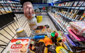 Ein Mann stellt ein Glas Joghurt in seinen Einkaufswagen im Supermarkt, in dem sich schon viele Lebensmittel nebeneinander und aufeinander befinden.