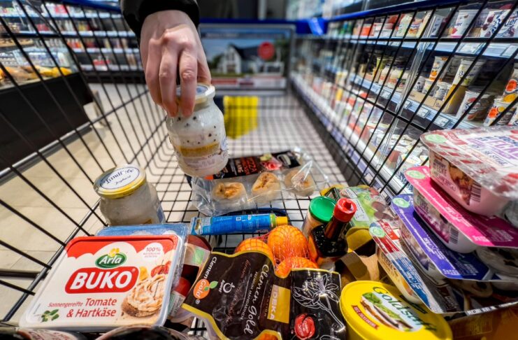 Ein Mann stellt ein Glas Joghurt in seinen Einkaufswagen im Supermarkt, in dem sich schon viele Lebensmittel nebeneinander und aufeinander befinden.