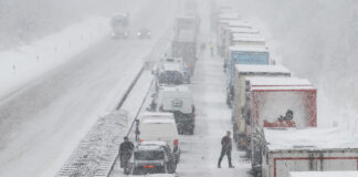 Es herrscht Stau auf einer Autobahn mitten im Winter. Die Straßen sind fast völlig eingeschneit und es wütet offenbar gerade ein weiterer Schneesturm. Die Lkws stehen dicht beieinander und haben ebenfalls eine Schicht Schnee auf ihren Dächern.