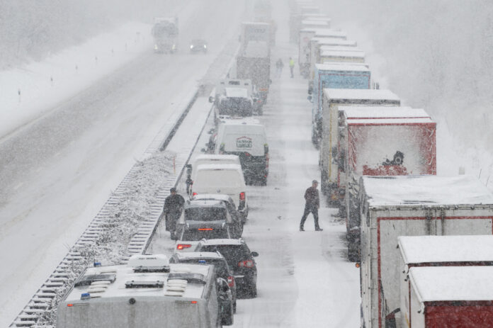 Es herrscht Stau auf einer Autobahn mitten im Winter. Die Straßen sind fast völlig eingeschneit und es wütet offenbar gerade ein weiterer Schneesturm. Die Lkws stehen dicht beieinander und haben ebenfalls eine Schicht Schnee auf ihren Dächern.