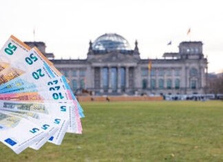 Einige Geldscheine werden wie ein Fächer vor dem Regierungsgebäude in die Luft gehalten. Mehrere unterschiedliche Euroscheine stehen dabei im Vordergrund.