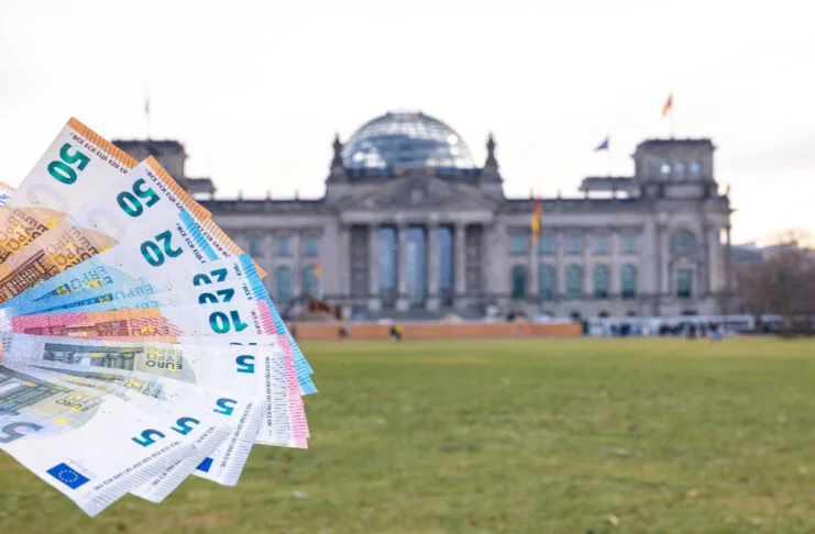 Einige Geldscheine werden wie ein Fächer vor dem deutschen Regierungsgebäude in Berlin in die Luft gehalten. Mehrere unterschiedliche Euro-Scheine sind dabei zu sehen, unter anderem 5-Euro-Scheine und 20-Euro-Scheine.