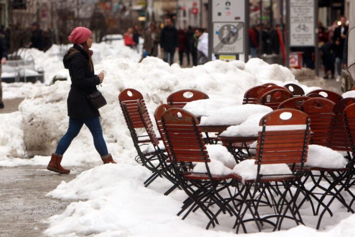 Eine Person in Wintersachen läuft durch eine Innenstadt im Winter mit sehr viel Schnee. Im Vordergrund Stühle und Tische im Freien, auf denen Schnee zu sehen ist. Im Hintergrund sind mehrere Menschen zu beobachten.