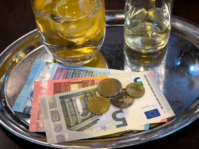 Getränke und Geld auf einem Tablett in einem Restaurant. Ein kleines und ein großes Glas mit Eiswürfeln stehen auf dem silbernen Tablett, auf dem auch ein 20-Euro-Schein, ein 10-Euro-Schein, ein 5-Euro-Schein sowie verschiedene Euro-Münzen liegen.