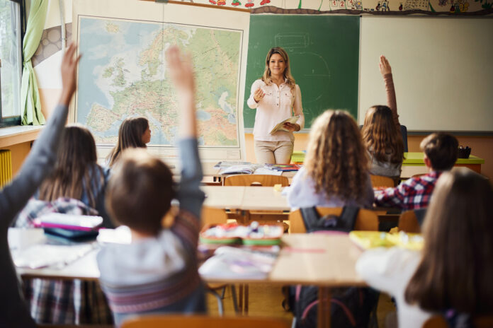 Eine Lehrerin steht im Klassenzimmer einer Schule vor vielen Schülern. Die Jungen und Mädchen sitzen an ihren Tischen und lernen, was ihnen im Unterricht beigebracht wird.