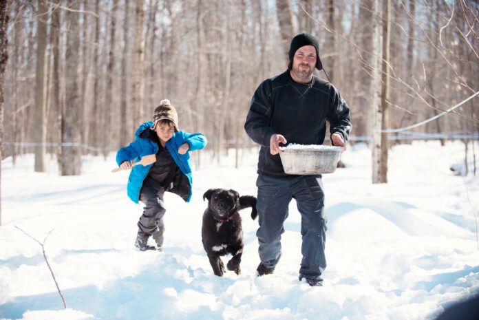 Ein Junge läuft mit seinem Vater und dem Hund durch den Schnee. Der Vater hält eine dampfende Schüssel in der Hand.