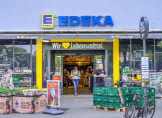 Der Eingang einer Filiale von Edeka mit zahlreichen Produkten am Eingang rechts und links. Das Logo sowie der Slogan des Supermarktes stehen im Bildfokus. Eine Frau verlässt den Laden.