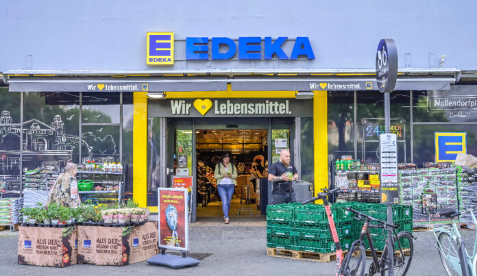 Der Eingang einer Filiale von Edeka mit zahlreichen Produkten am Eingang rechts und links. Das Logo sowie der Slogan des Supermarktes stehen im Bildfokus. Eine Frau verlässt den Laden.
