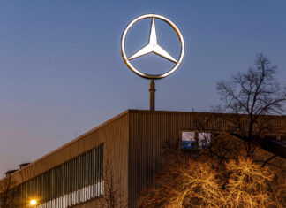 Ein großer Mercedes-Stern glänzt als Daimler-Logo von Mercedes prägt auf dem riesigen Firmengebäude. Im Lichterschein ist im Vordergrund ein Baum zu erkennen. Im Hintergrund geht gerade die Sonne unter.