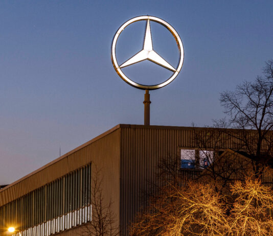 Ein großer Mercedes-Stern glänzt als Daimler-Logo von Mercedes prägt auf dem riesigen Firmengebäude. Im Lichterschein ist im Vordergrund ein Baum zu erkennen. Im Hintergrund geht gerade die Sonne unter.
