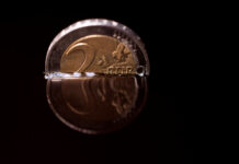 Eine 2-Euro-Münze in schwarz. Sie ist in eine Flüssigkeit getaucht und nur die obere Hälfte schaut heraus. Der Hintergrund ist komplett in Schwarz gehalten.