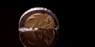 Eine 2-Euro-Münze in schwarz. Sie ist in eine Flüssigkeit getaucht und nur die obere Hälfte schaut heraus. Der Hintergrund ist komplett in Schwarz gehalten.