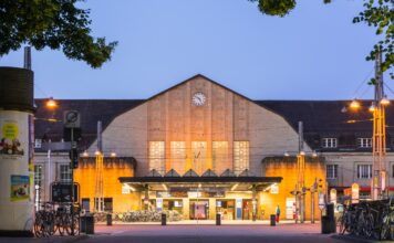 Der Hauptbahnhof Karlsruhe bei Sonnenaufgang. Die große beleuchtete Fassade des Hauptbahnhofs mit der Uhr ist zu sehen. Der Eingangsbereich ist ebenso beleuchtet und wirkt auf den ersten Blick einladend auf die Reisenden.