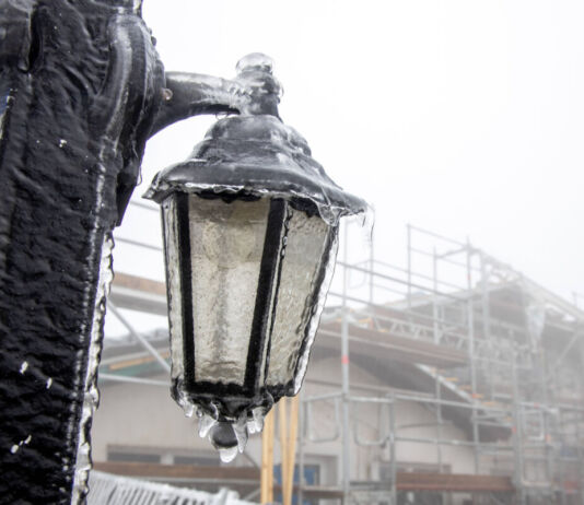 Unter grauem Himmel ist ein Lichtmast durch Schnee- und Eisregen eingefroren. Im Hintergrund ist ein ebenso gefrorenes Baugerüst zu sehen.