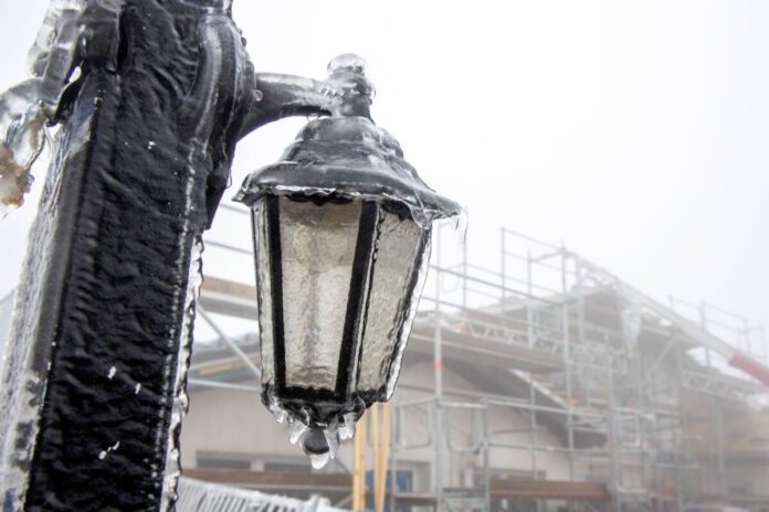 Unter grauem Himmel ist ein Lichtmast durch Schnee- und Eisregen eingefroren. Im Hintergrund ist ein ebenso gefrorenes Baugerüst zu sehen.