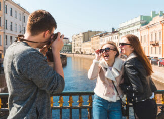Ein Mann fotografiert 2 lachende Frauen bei einem Fotoshooting auf einer Brücke vor einer pittoresken Stadtkulisse im Sonnenschein.