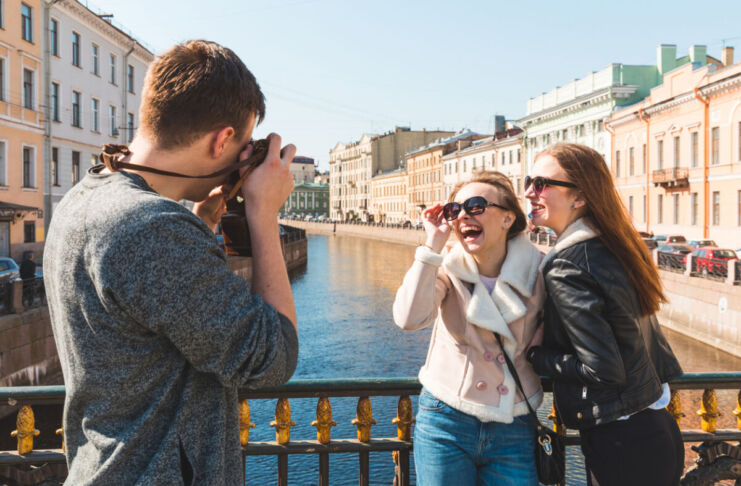 Ein Mann fotografiert 2 lachende Frauen bei einem Fotoshooting auf einer Brücke vor einer pittoresken Stadtkulisse im Sonnenschein.