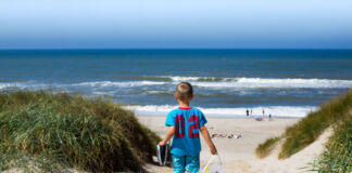 Ein Junge im blauen Shirt mit Schuhen in seiner Hand spaziert an einem Sommertag barfuß am Strand, während im Vordergrund Strandbesucher und Schwimmer im Meer zu sehen sind.