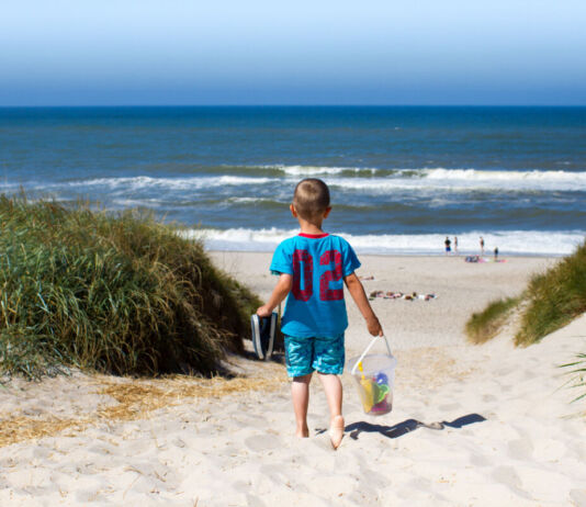 Ein Junge im blauen Shirt mit Schuhen in seiner Hand spaziert an einem Sommertag barfuß am Strand, während im Vordergrund Strandbesucher und Schwimmer im Meer zu sehen sind.