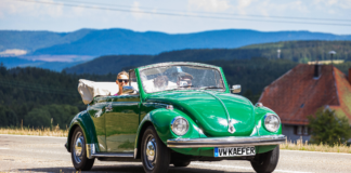 Ein VW-Käfer, der inzwischen ein wahrer Oldtimer ist, fährt auf einer Landstraße. Im Hintergrund befinden sich Berge.