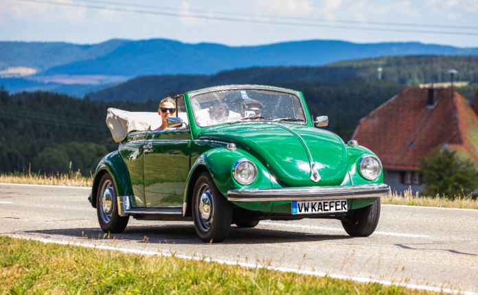 Ein VW-Käfer, der inzwischen ein wahrer Oldtimer ist, fährt auf einer Landstraße. Im Hintergrund befinden sich Berge.