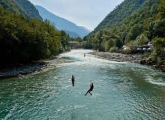 Mehrere Touristen betreiben Ziplining an einem wilden, schlängelnden Fluss bei bestem Wetter in einer bergigen Landschaft. Im Hintergrund sind noch mehr Berge und ein blauer Himmel zu sehen.
