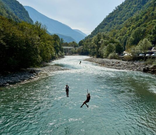 Mehrere Touristen betreiben Ziplining an einem wilden, schlängelnden Fluss bei bestem Wetter in einer bergigen Landschaft. Im Hintergrund sind noch mehr Berge und ein blauer Himmel zu sehen.