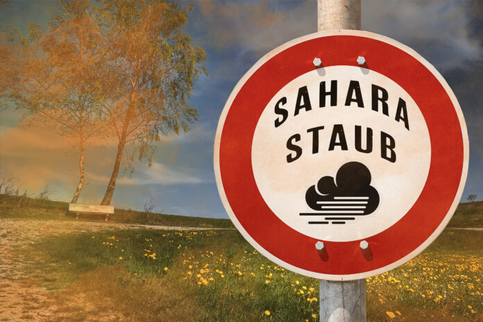 Ein rundes Schild auf dem eine Warnung vor Sahara-Staub steht. Im Hintergrund erkennt man eine Wiese und Wüstenstaub der darüber hinweg weht.