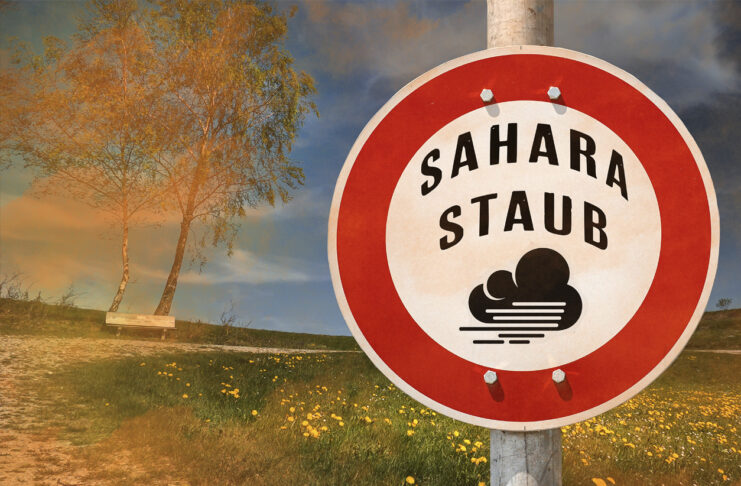 Ein rundes Schild auf dem eine Warnung vor Sahara-Staub steht. Im Hintergrund erkennt man eine Wiese und Wüstenstaub der darüber hinweg weht.