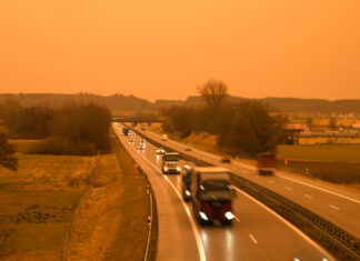 Der Blick auf eine Autobahn während eines Blutregens oder aber eines Sonnenaufgangs oder Sonnenuntergangs. Man sieht mehrere LKWs hintereinander in einer Reihe fahren. Sie haben ihre Scheinwerfer bereits an.