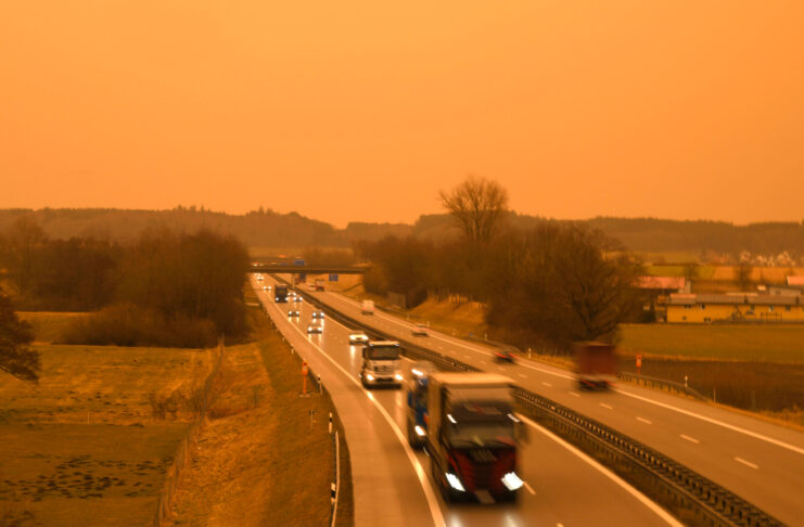 Der Blick auf eine Autobahn während eines Blutregens oder aber eines Sonnenaufgangs oder Sonnenuntergangs. Man sieht mehrere LKWs hintereinander in einer Reihe fahren. Sie haben ihre Scheinwerfer bereits an.
