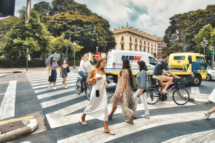 In einer dicht gedrängten Altstadt einer Innenstadt überqueren viele Menschen eine Straße über einen Zebrastreifen. Autos, Fahrradfahrer und Passanten sind gleichzeitig und sicher unterwegs.